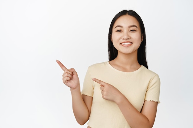 Gros plan d'une fille coréenne souriante pointant les doigts vers la gauche montrant une publicité et ayant l'air heureux de donner des informations sur fond blanc