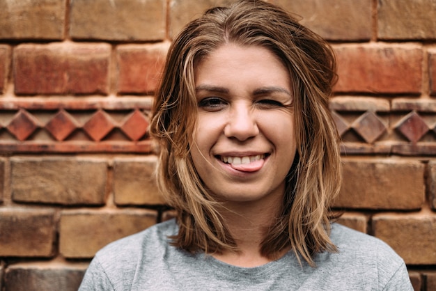 Gros plan d'une fille avec une coupe courte et des cheveux blonds montre en plaisantant sa langue contre un mur de briques