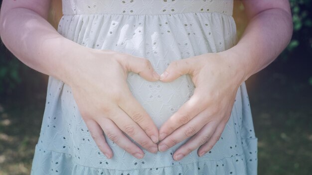 Gros plan d'une fille enceinte vêtue d'une robe bleue douce avec les mains pliées en forme de coeur dans le