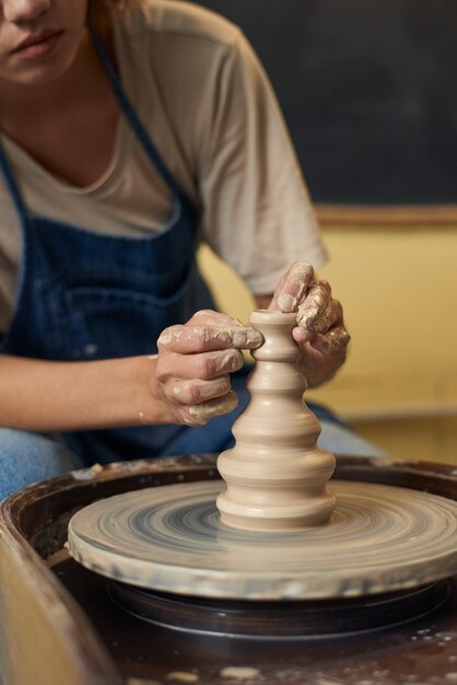 Gros plan d'une fille méconnaissable en tablier sculptant un beau vase en argile avec des mains sales sur un tour de potier