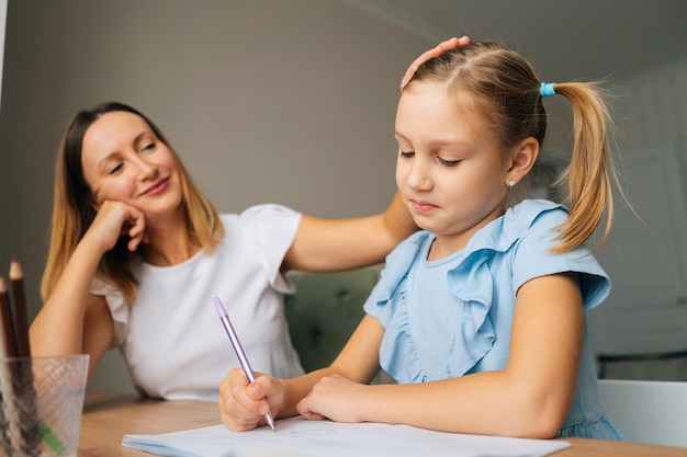 Gros plan d'une fille primaire mignonne faisant ses devoirs avec une jeune mère assise à la table de la maison par la fenêtre