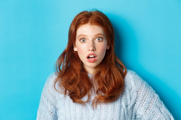 Gros plan d'une fille rousse surprise qui regarde la caméra sans voix, bouche ouverte avec admiration, debout sur fond bleu