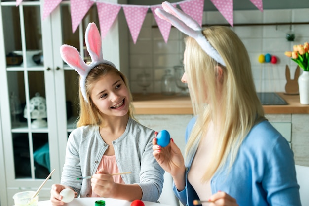 Gros plan d'une fille souriante regardant maman qui peint des oeufs de Pâques.