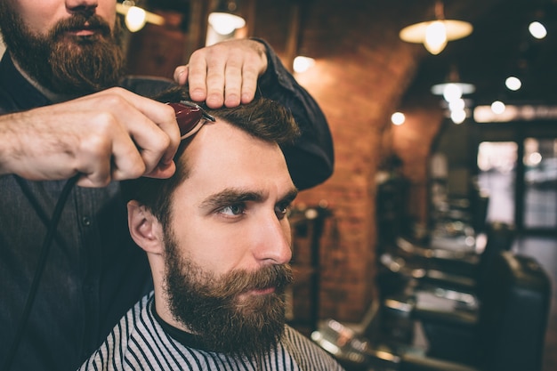 Gros plan d'un gars barbu. Son coiffeur coupe et modèle ses cheveux.