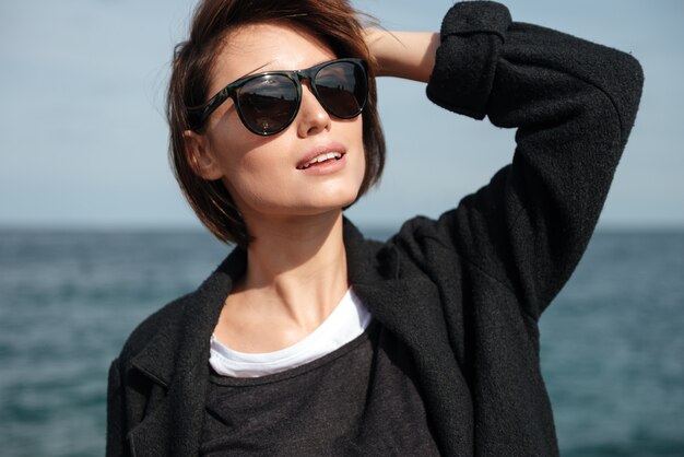 Gros plan de l'heureuse belle jeune femme à lunettes de soleil debout au bord de la mer