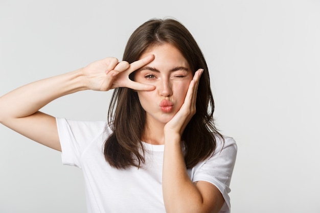 Gros plan de l'heureuse jolie fille brune clignotant idiot et montrant le geste de paix kawaii, blanc.