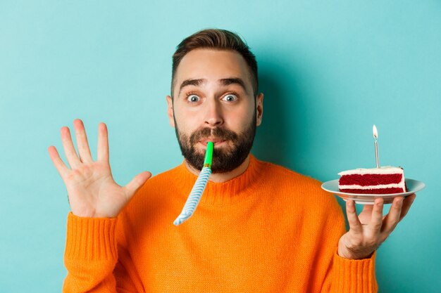 Gros plan d'un homme adulte drôle célébrant son anniversaire, tenant un gâteau bday avec bougie, soufflant le sifflet de fête et se réjouissant, debout sur fond bleu clair.