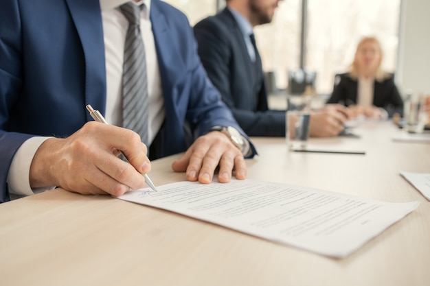 Gros plan d'un homme d'affaires méconnaissable en costume assis à table et signant un contrat lors d'une réunion avec des partenaires commerciaux