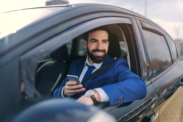 Gros plan d'homme d'affaires souriant à l'aide de téléphone intelligent tout en étant assis dans sa voiture et en s'appuyant sur la fenêtre.