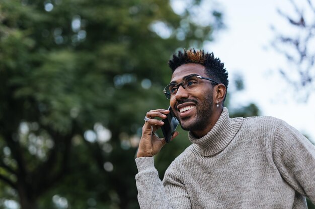 Gros plan sur un homme afro-américain souriant en gilet à col roulé assis sur un banc de parc et parlant sur son téléphone portable