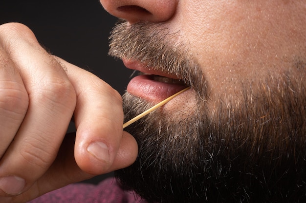 Gros plan homme barbu nettoyer les dents de la nourriture coincé ses dents avec un cure-dent après le petit déjeuner déjeuner dîner...