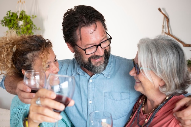 Gros plan d'un homme barbu souriant embrassant sa femme et sa mère ensemble s'amusant et portant un toast avec un verre de vin rouge dans la chambre domestique