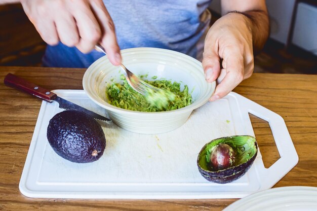 Gros plan homme caucasien assis dans sa cuisine préparant du guacamole avec une fourchette dans un bol blanc