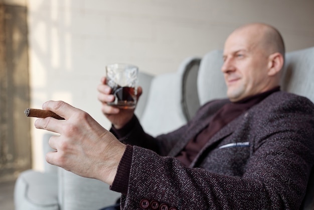 Gros plan d'un homme chauve détendu en veste élégante assis dans un fauteuil et fumer un cigare tout en buvant du whisky