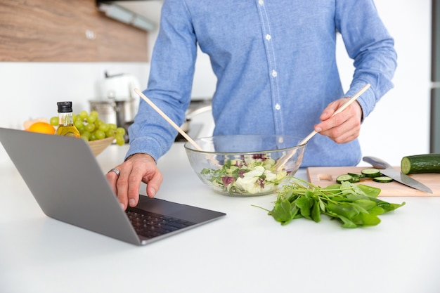 Gros plan de l'homme en chemise bleue utilisant un ordinateur portable et faisant de la salade dans un bol en verre sur la cuisine