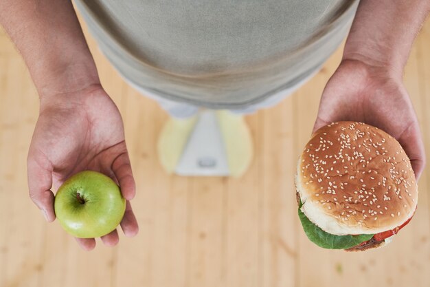 Gros plan d'un homme debout sur une balance tenant une pomme dans une main et un hamburger dans l'autre pour faire un d...