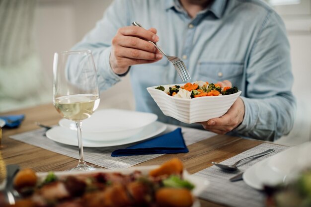 Gros plan d'un homme méconnaissable prenant de la salade dans un bol pendant le déjeuner à la table à manger