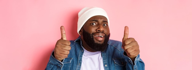 Photo gros plan d'un homme noir maladroit en bonnet montrant le pouce levé mais se sentant grincer des dents debout incertain et inquiet