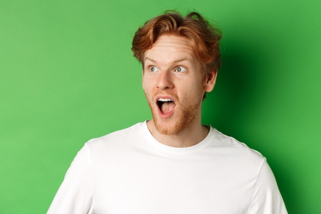 Gros plan d'un homme rousse surpris et impressionné par l'offre de promotion, regardant à gauche avec la mâchoire tombée, debout sur fond vert.