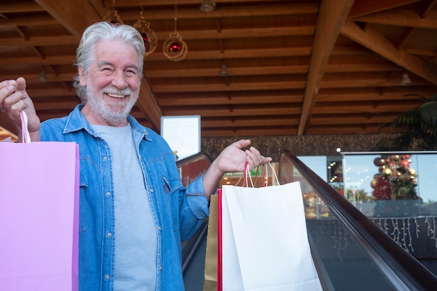 Gros plan sur un homme senior souriant aux cheveux blancs faisant du shopping pour Noël dans un centre commercial, regardant la caméra tout en tenant de nombreux sacs avec des cadeaux pour la famille et les amis