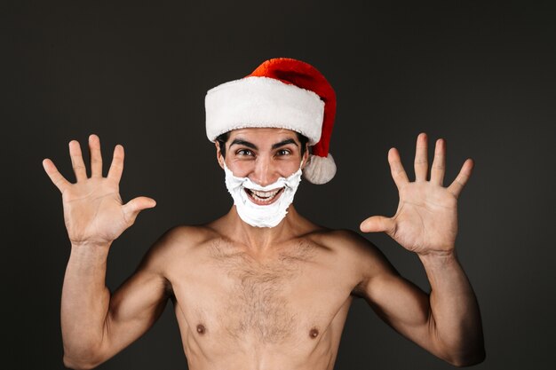 Photo gros plan d'un homme torse nu excité portant chapeau de père noël debout isolé, avec les mains levées