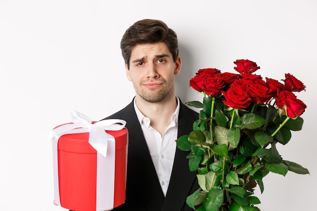 Gros plan d'un homme triste en costume, tenant un bouquet de roses rouges et un cadeau, debout contrarié sur fond blanc