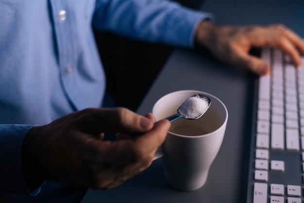 Gros plan sur un homme versant du sucre sur une tasse de café avec une cuillère en métal travaillant sur ordinateur tard dans la nuit