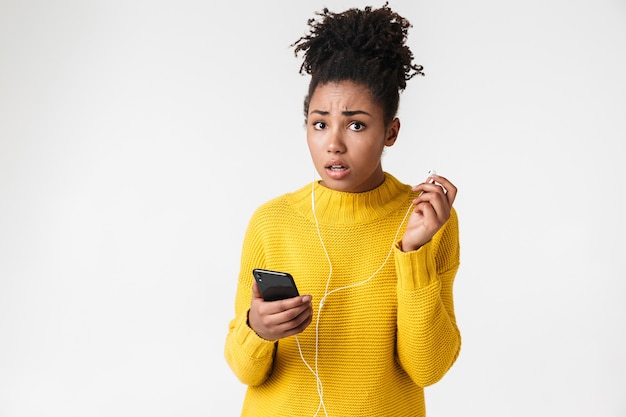 Gros plan de l'image de la femme afro-américaine perplexe tenant le téléphone portable et écouter de la musique avec des écouteurs sur blanc