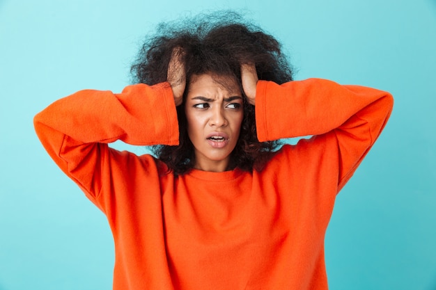 Gros plan de l'image de la femme confuse en chemise rouge à côté et saisissant la tête avec des cheveux hirsutes, isolé sur mur bleu