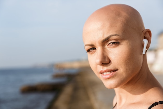 Gros plan de l'image d'une jolie femme chauve à l'aide d'écouteurs près de la mer en matinée ensoleillée