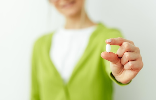 Gros plan image d'une pilule à la main avec vue floue d'une femme souriante Belle jeune femme portant un cardigan vert et un t-shirt blanc prenant la pilule isolé sur un mur blanc