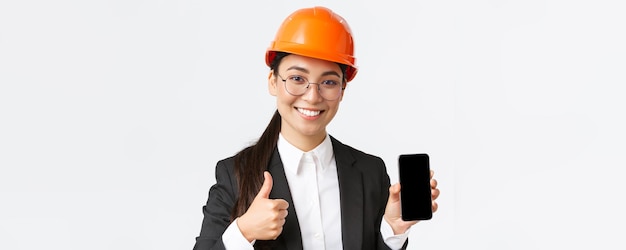 Gros plan d'une ingénieure professionnelle souriante responsable de la construction en costume d'affaires et casque de sécurité montrant l'écran du smartphone et le pouce levé en approbation fond blanc