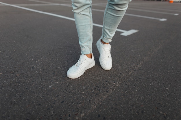 Gros plan des jambes mâles. Tendance jeune homme à la mode en jeans bleu vintage en cuir élégantes baskets blanches se tiennent sur la route goudronnée à l'extérieur. Style de rue moderne pour les jeunes.