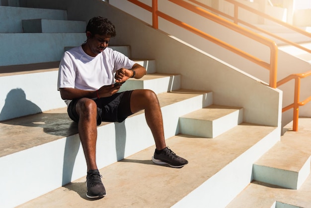 Gros plan sur un jeune athlète asiatique coureur de sport homme noir porter une montre intelligente de l'heure moderne et se synchroniser avec un smartphone vérifier le résultat de la course après une course d'entraînement à l'extérieur, technologie d'entraînement d'exercice sain