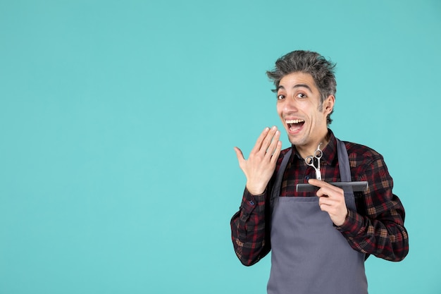 Gros plan d'un jeune coiffeur souriant et heureux portant un tablier gris et tenant un peigne et des ciseaux sur fond de couleur bleu