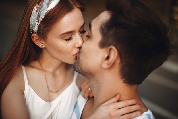 Gros plan d'un jeune couple aimant s'embrasser les yeux fermés. Charmante jeune femme aux cheveux roux et aux taches de rousseur embrassant son petit ami en plein air lors de rencontres.