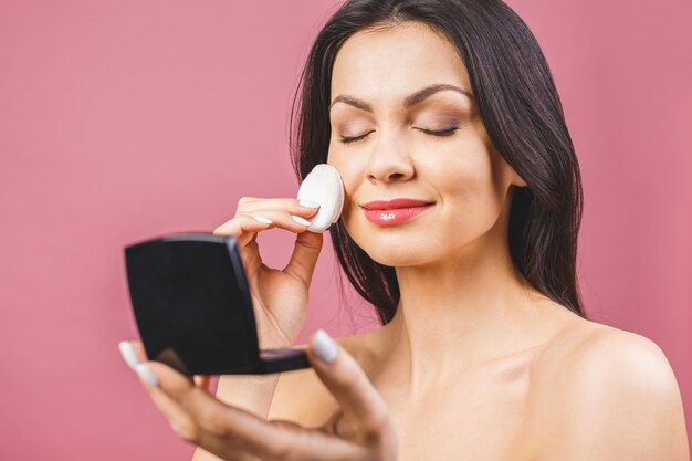 Gros plan jeune femme appliquant la poudre sur le visage isolé sur fond rose.