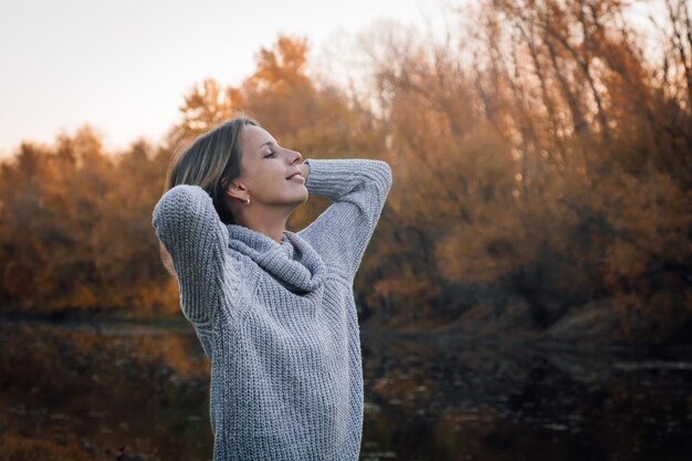 Gros plan d'une jeune femme caucasienne blonde debout dans la forêt regardant vers le haut souriant avec les deux mains derrière la tête avec des arbres couverts de coucher de soleil en arrière-plan Promenade d'automne dans la forêt