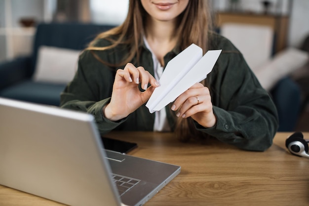 Gros plan d'une jeune femme faisant un avion en papier assis à table avec un ordinateur portable et des documents