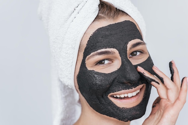 Gros plan d'une jeune femme heureuse souriante avec des dents blanches appliquant un masque organique d'argile noire cosmétique faciale sur le visage porte une serviette blanche sur les cheveux La femme prend soin de sa peau isolée sur un mur blanc