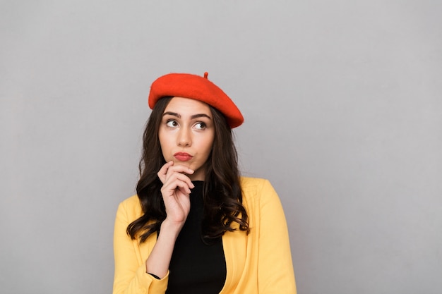 Gros plan d'une jeune femme pensive au chapeau rouge debout sur fond gris, à l'écart