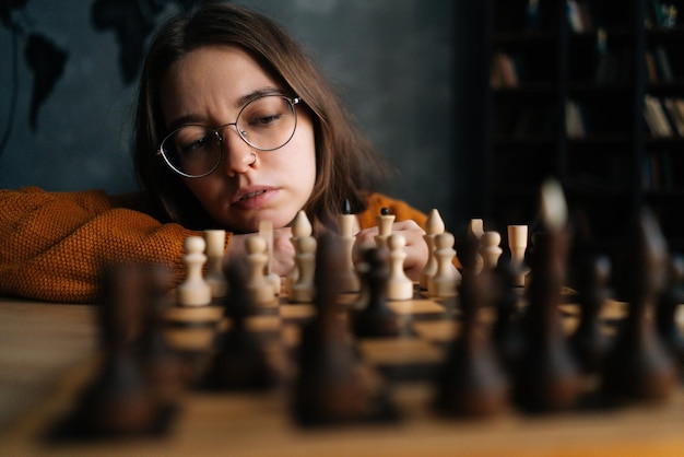 Gros plan d'une jeune femme pensive dans des lunettes élégantes pensant au mouvement d'échecs assis sur le sol dans une pièce sombre mise au point sélective