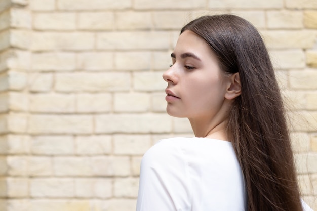 Gros plan d'une jeune fille brune en T-shirt blanc aux cheveux longs regarde sur le côté sur fond de mur de briques jaunes. place pour votre conception