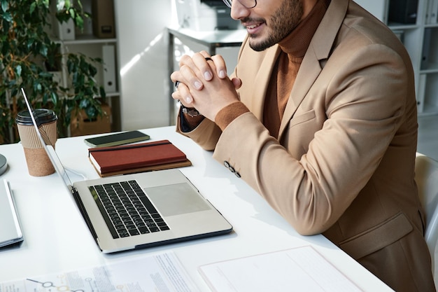 Gros plan d'un jeune homme d'affaires en veste assis à table et utilisant une application de vidéoconférence tout en communiquant en ligne au bureau