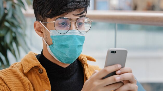 Gros plan jeune homme arabe dans un masque de protection en tapant un message sur un smartphone à l'aide d'une application mobile