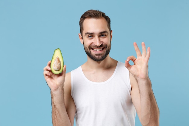 Gros plan sur un jeune homme barbu de 20 ans en chemise blanche tenant la moitié de l'avocat isolé sur fond bleu pastel, portrait en studio. Concept de procédures cosmétiques de soins de la peau. Maquette de l'espace de copie.