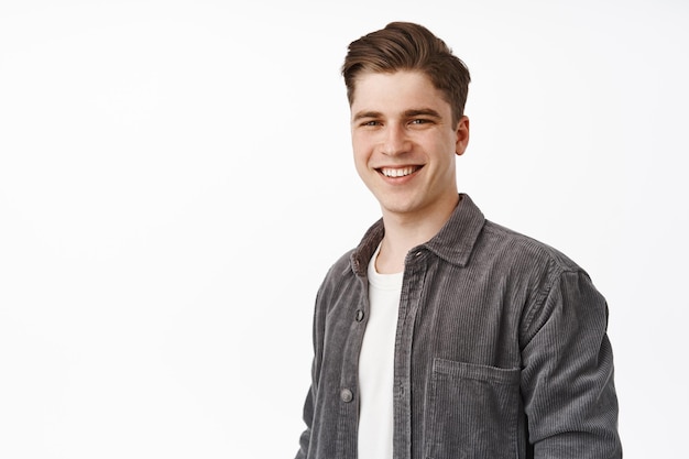 Gros plan d'un jeune homme candide, bel homme souriant aux dents blanches, debout détendu et décontracté sur fond de studio isolé, l'air heureux, fond blanc