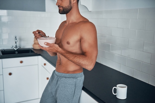 Gros plan sur un jeune homme torse nu prenant son petit-déjeuner en se tenant debout dans la cuisine à domicile