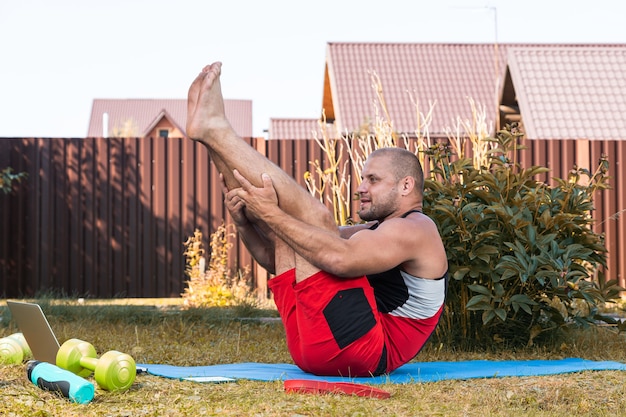 Gros plan sur un jeune homme en uniforme de sport essayant de méditer dans une position difficile, s'étirant sur le sol dans l'arrière-cour en été, disons