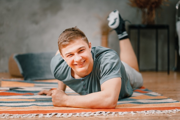 Gros plan d'un jeune homme en uniforme de sport repose sur le sol à la maison, regardant la caméra et souriant sur la chambre. Étudiant flânant et retardant le travail et les études